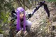 A woman with purple hair and a scythe in the woods.